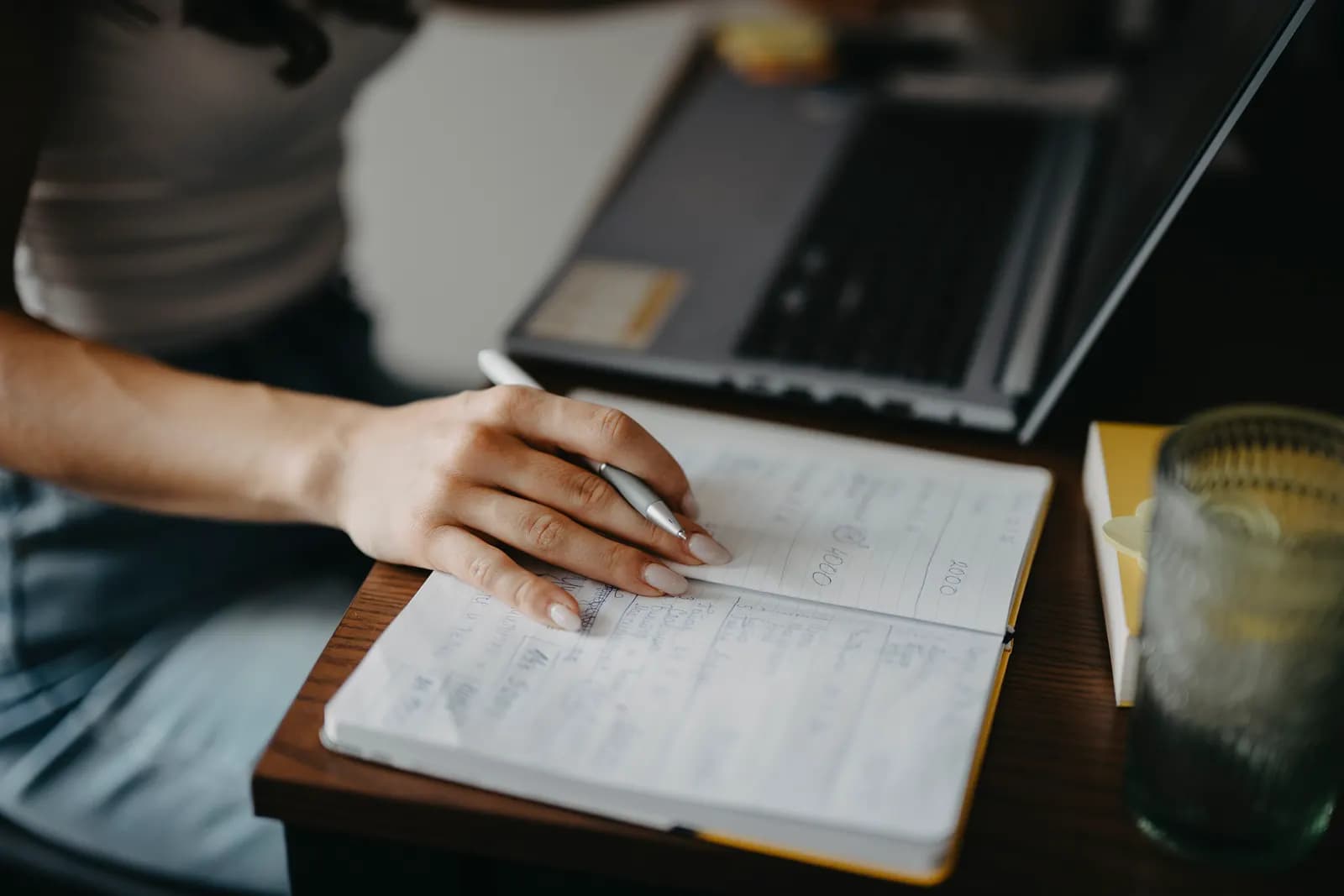 A person writing notes in a notebook at a wooden desk beside an open laptop