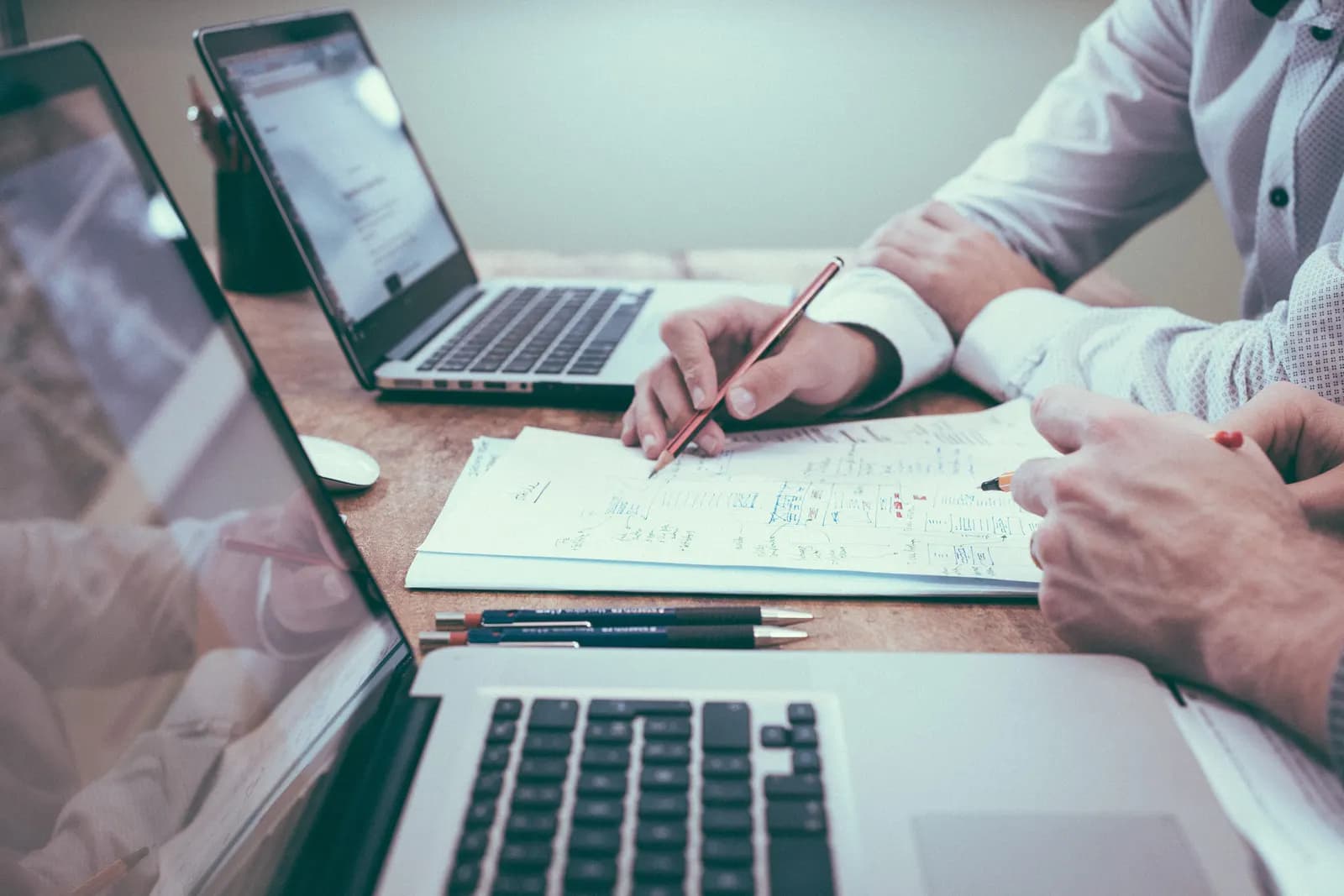 Two people at a wooden table with open laptops, one marking up printed notes with a pencil