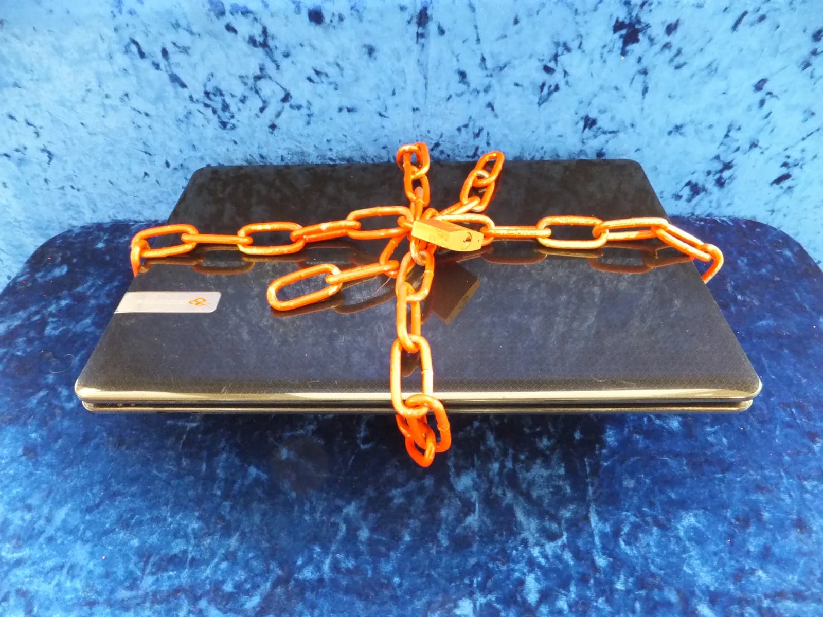 Closed laptop wrapped in an orange chain and padlock on a blue textured background