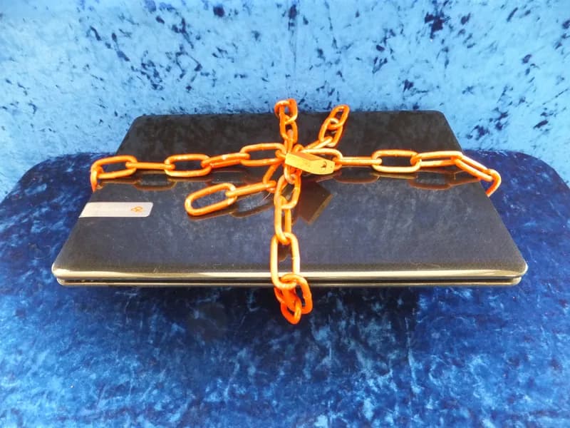 Closed laptop wrapped in an orange chain and padlock on a blue textured background
