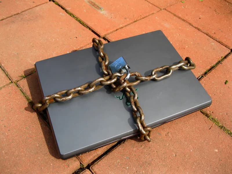 Closed grey laptop wrapped in a rusty chain fastened with a padlock, lying on red paving bricks