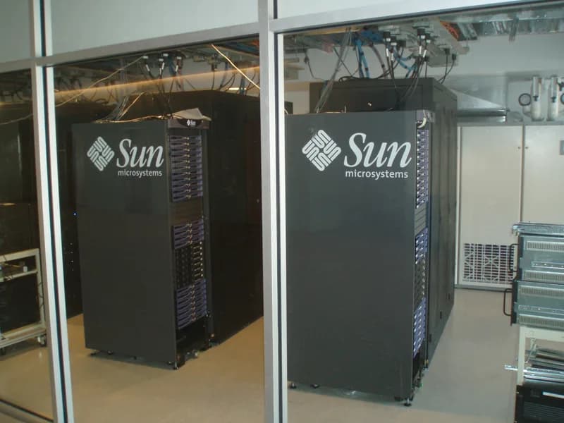 Two black Sun Microsystems server racks with cables running from their tops in a glass-walled server room