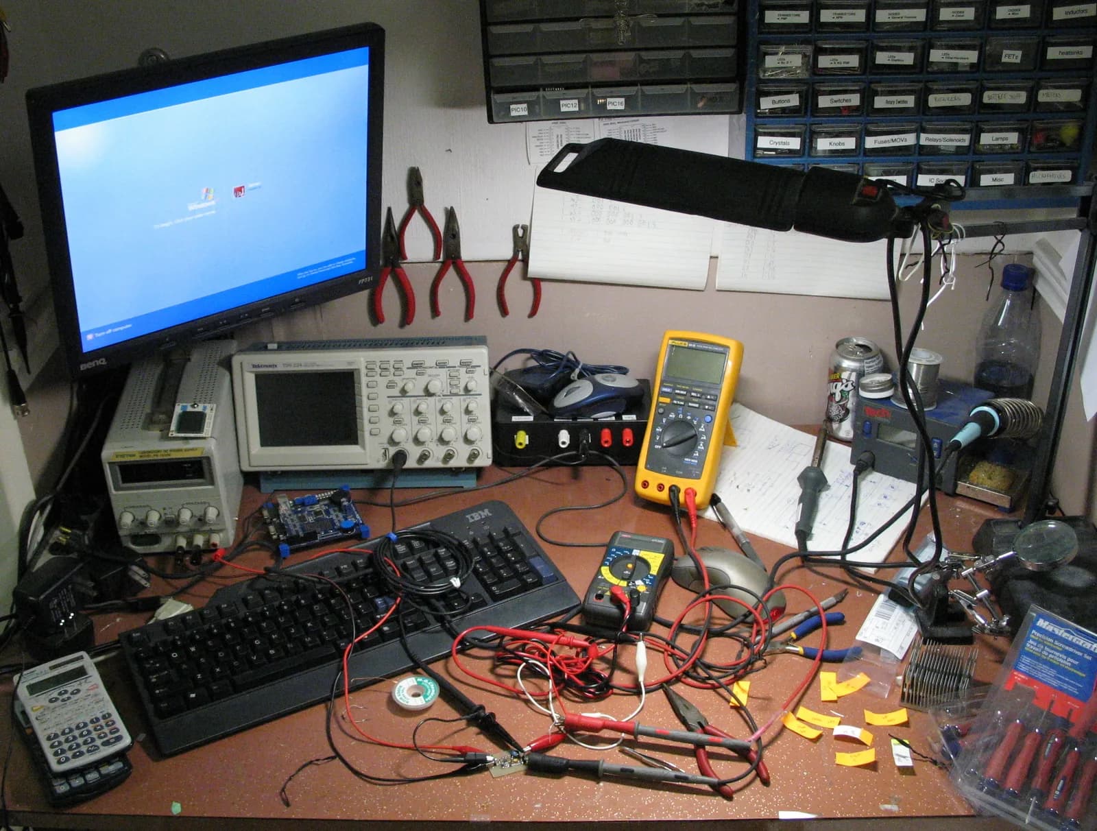 Cluttered electronics workbench with an oscilloscope, two multimeters, a power supply, test leads and a computer monitor