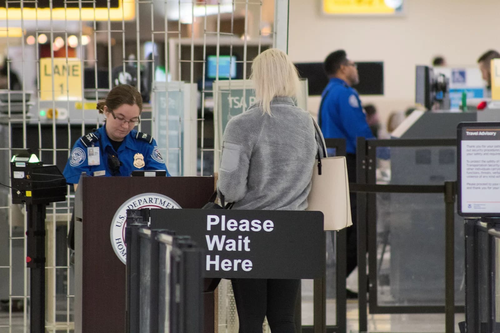 Airport security officer at a podium checking a passenger’s identification beside a Please Wait Here sign