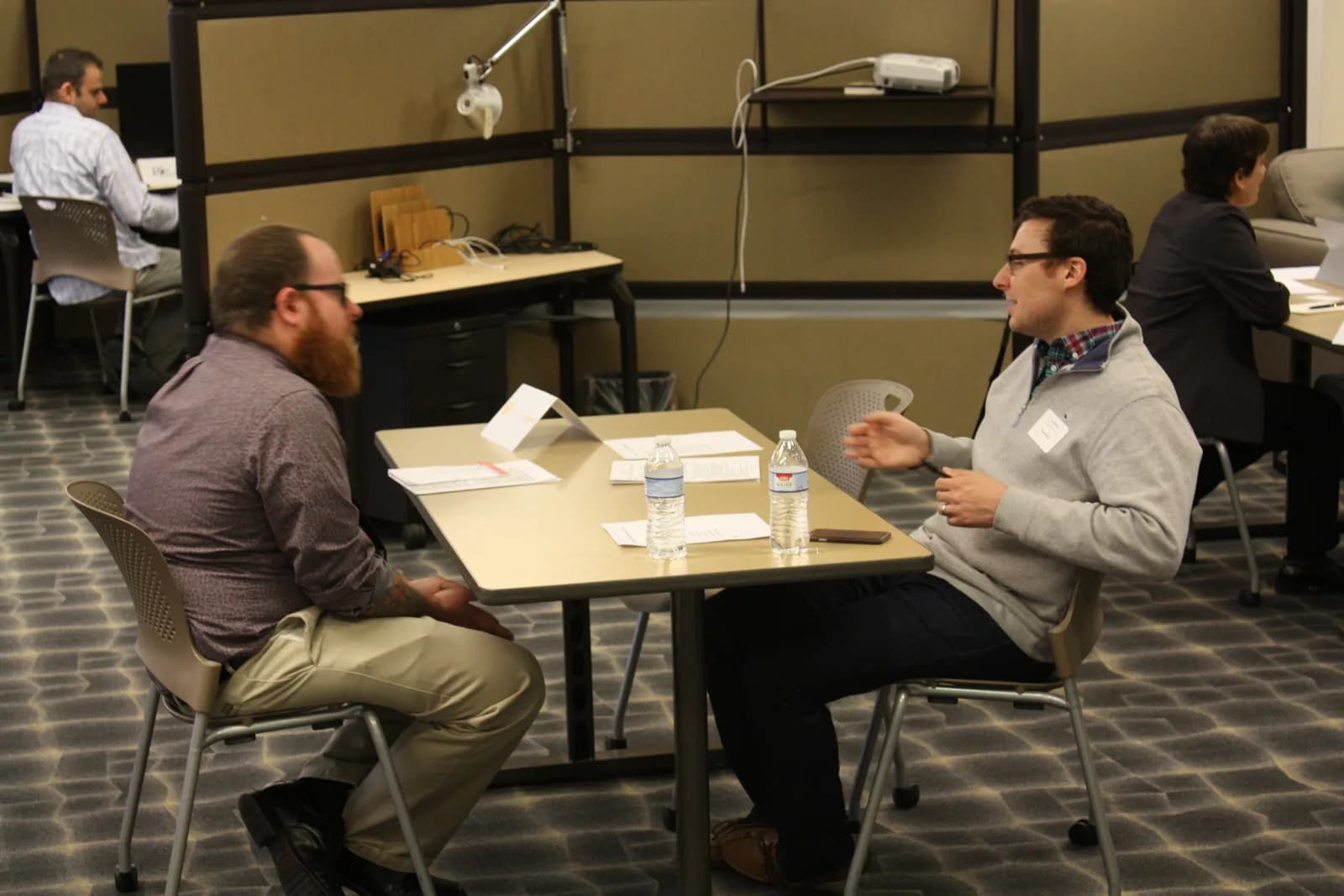 Two men talking across a small table during a technical job interview session, with water bottles and printed résumés on the table