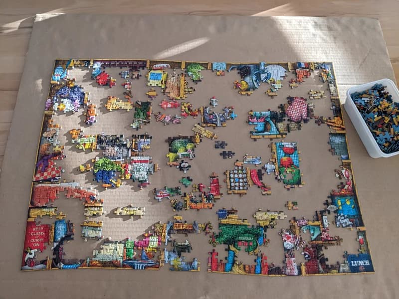 Partly assembled jigsaw puzzle on a cardboard sheet with a box of loose pieces beside it