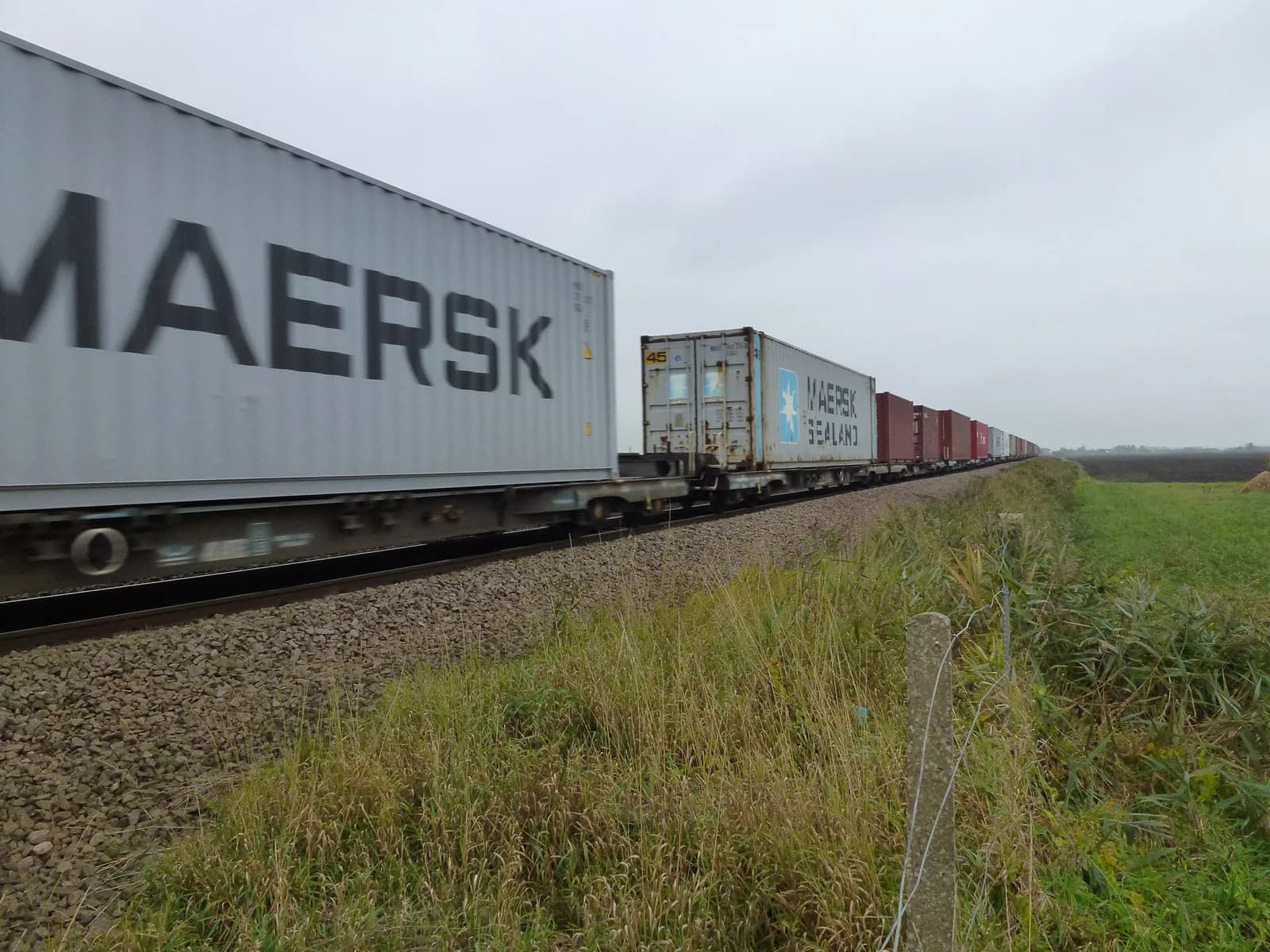 Long freight train of shipping containers on flat wagons passing through flat countryside