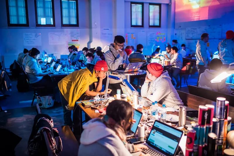 A crowded hackathon hall lit in blue where developers work on laptops at long shared tables