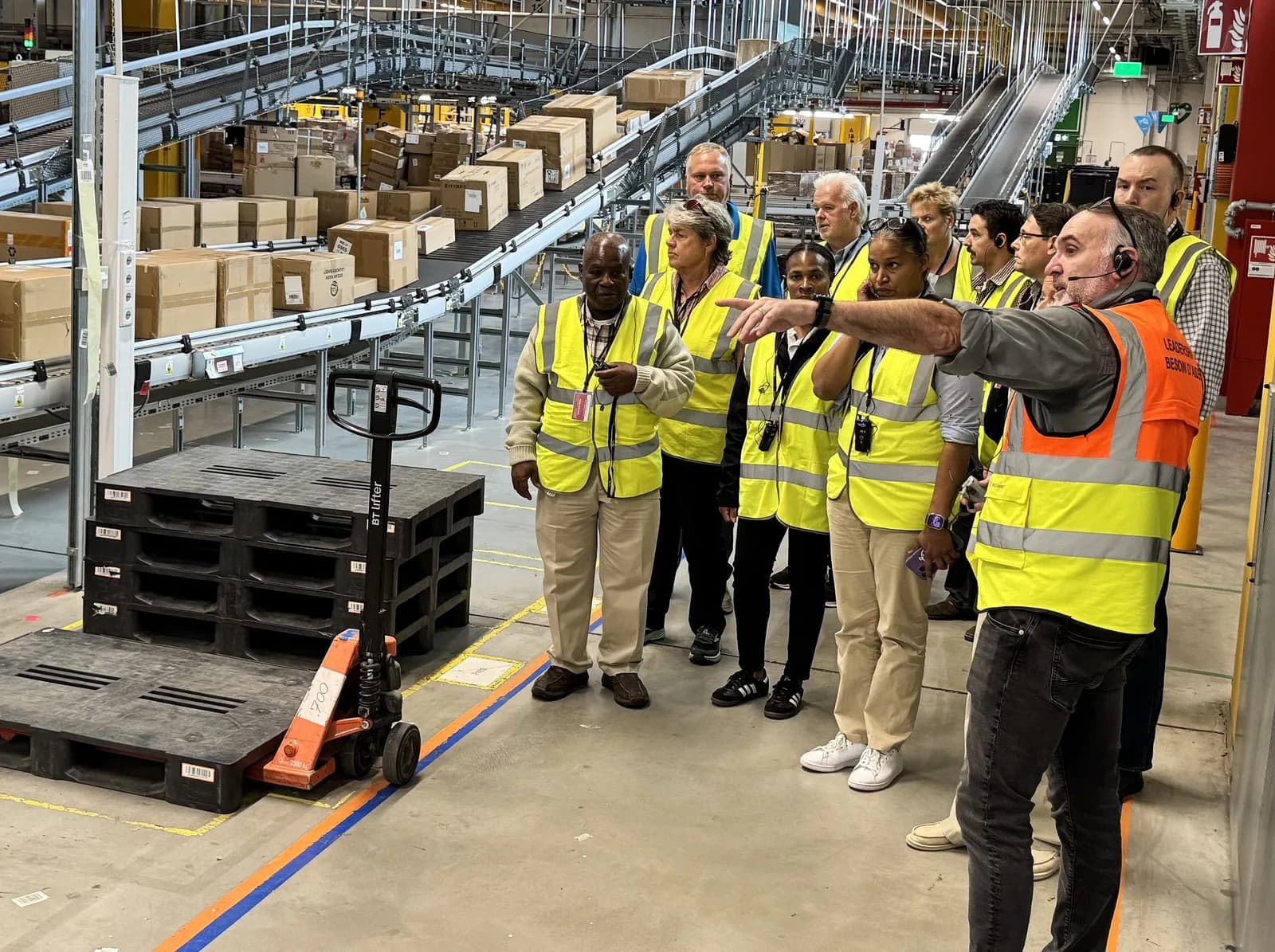 Visitors in high-visibility vests listen to a guide beside conveyor belts and stacked boxes in a fulfilment centre