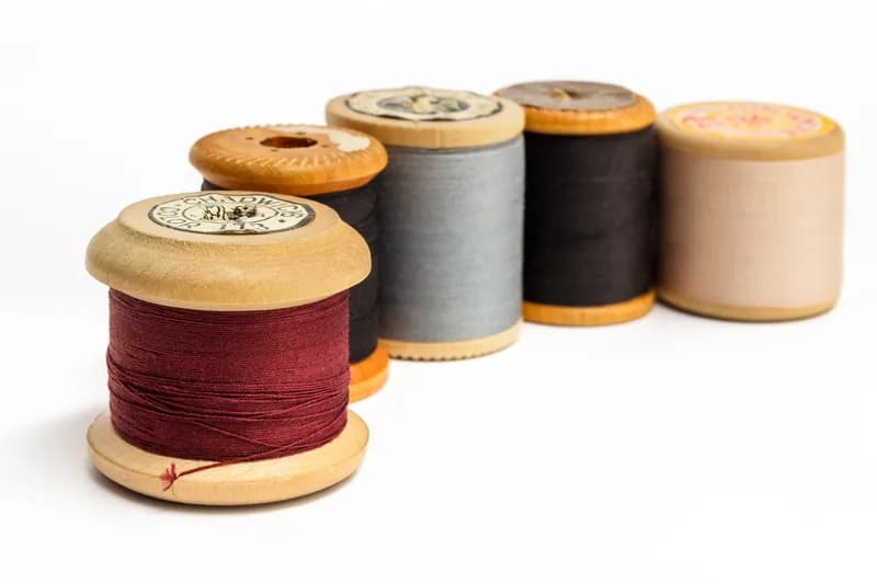 Five old wooden spools of cotton sewing thread in red, black, grey, dark brown and beige lined up on a white background