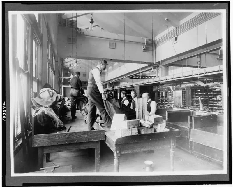Early 20th-century black-and-white photo of postal workers emptying mail sacks onto wooden tables in a Washington, D.C. sorting room
