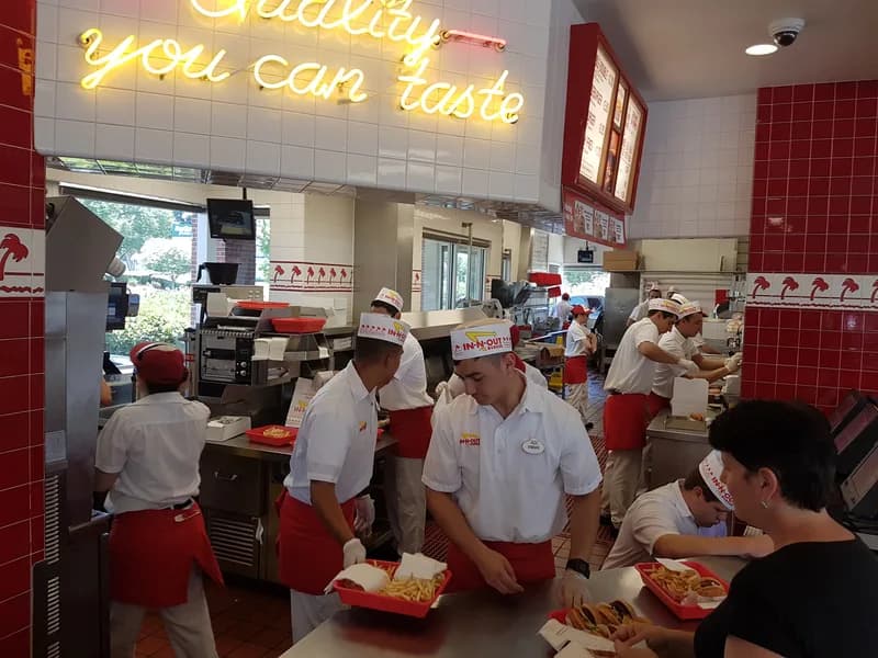 Busy fast-food kitchen with staff in white uniforms and red aprons preparing orders at a counter