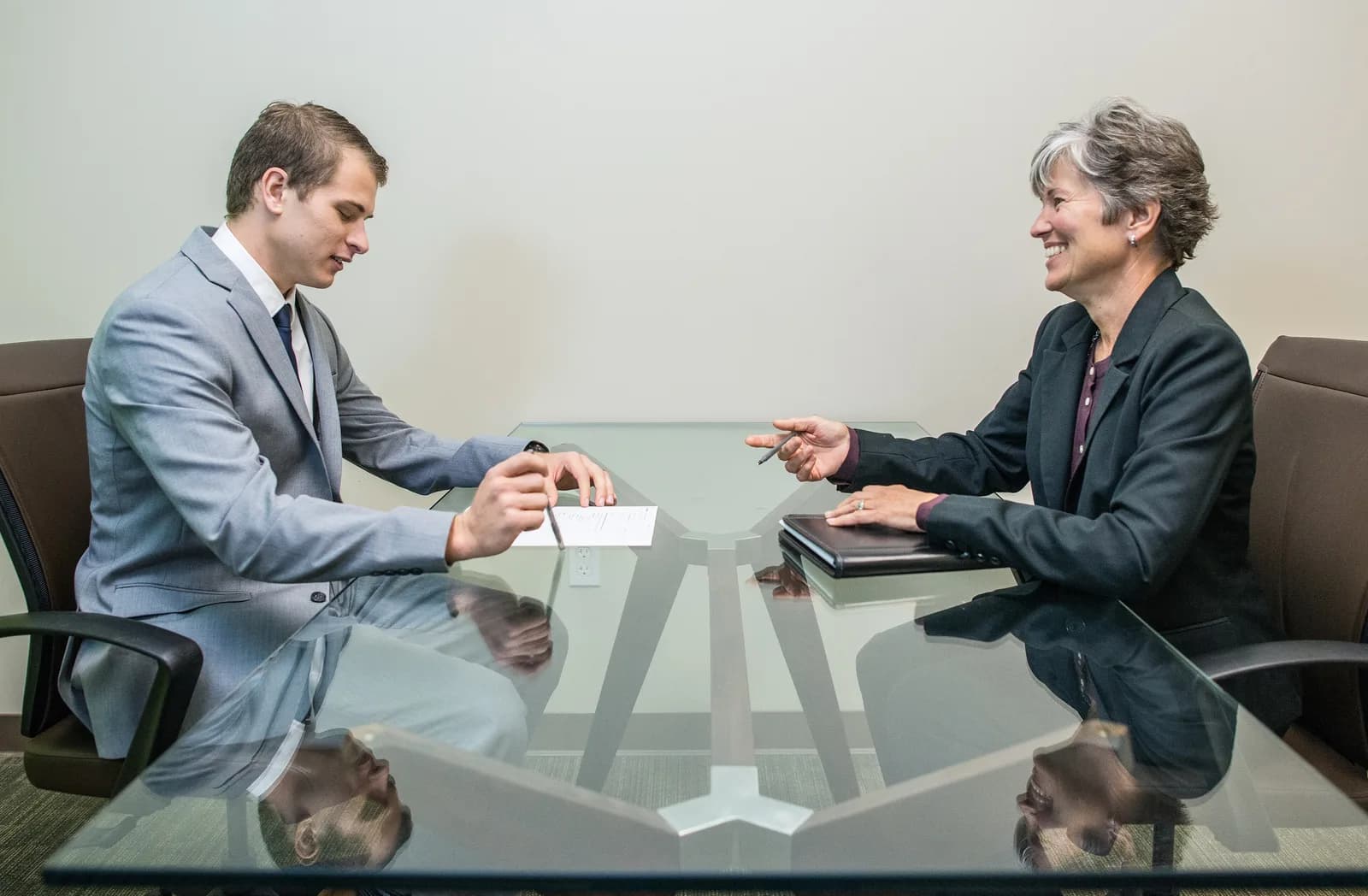 Young man in a grey suit writing on a sheet of paper at a glass table while a smiling interviewer talks