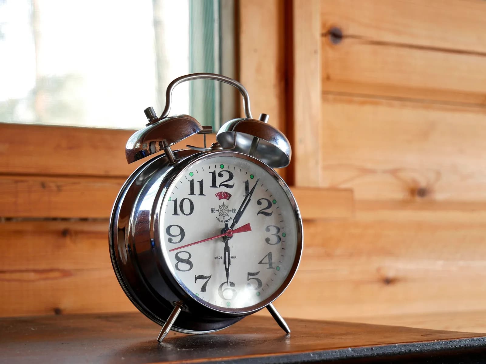 A chrome twin-bell mechanical alarm clock on a wooden table in front of a window and a log wall