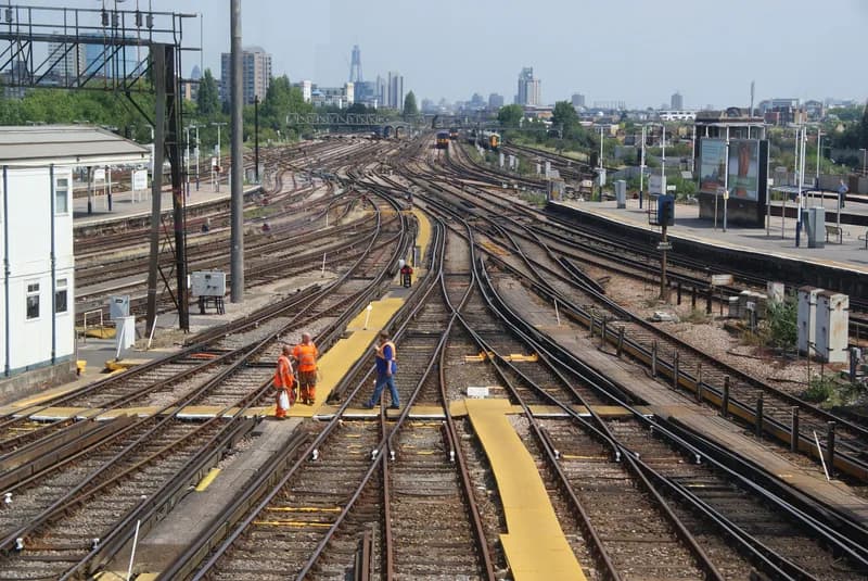 Many railway tracks and sets of points converging at Clapham Junction, with track workers in orange high-visibility clothing and the London skyline behind
