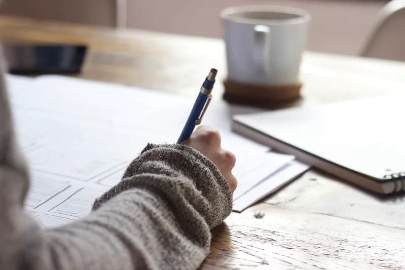Hand holding a pen over printed wireframe sketches on a wooden table, next to a coffee mug and notebook