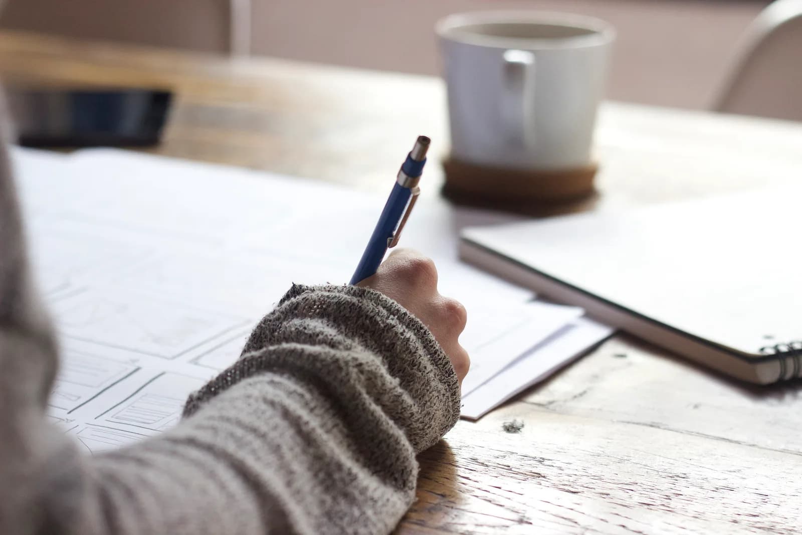 Hand holding a pen over printed wireframe sketches on a wooden table, next to a coffee mug and notebook