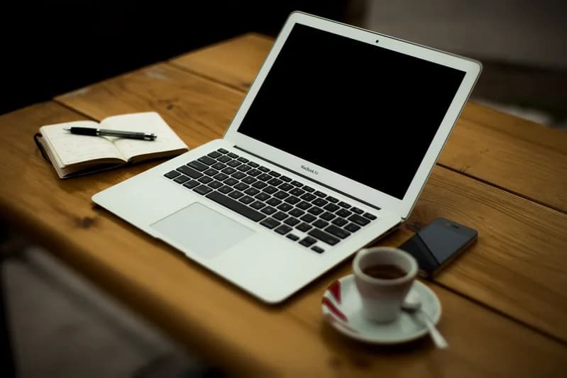Laptop on a wooden table beside an open notebook with a pen, a smartphone and an espresso cup
