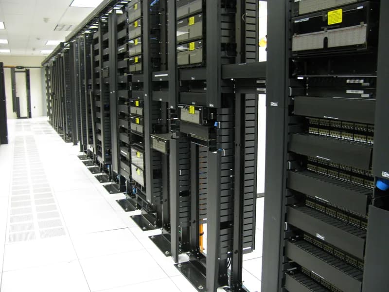 Aisle of black network racks with patch panels and cable managers on a white raised floor in a data centre