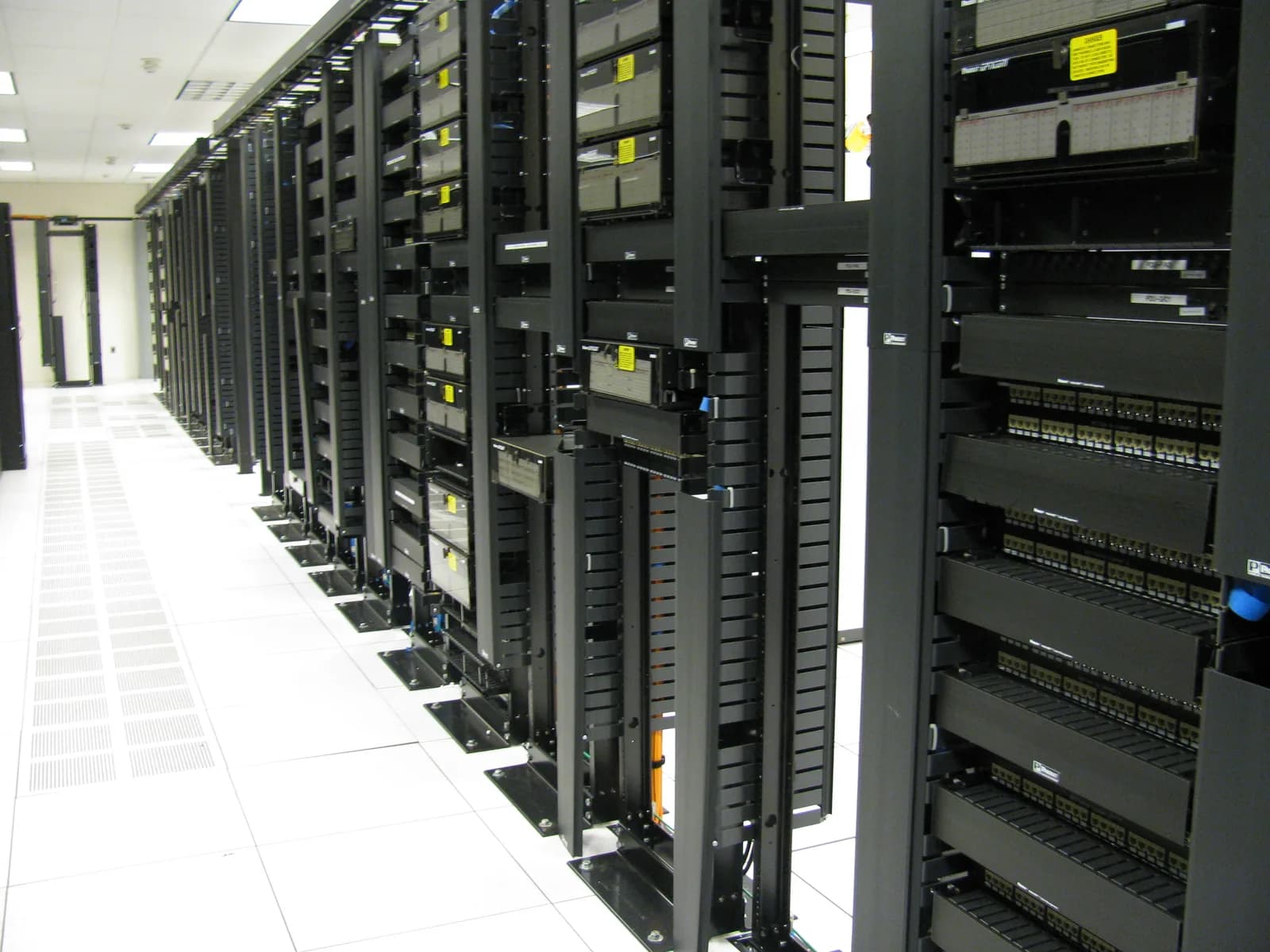 Aisle of black network racks with patch panels and cable managers on a white raised floor in a data centre