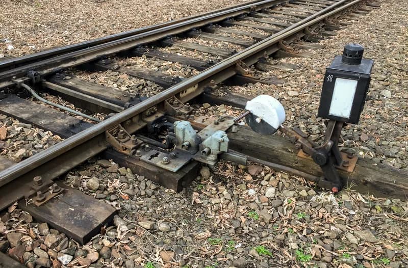 Hand-operated railway switch with a white counterweight lever and a lamp box beside the rails on a gravel track bed
