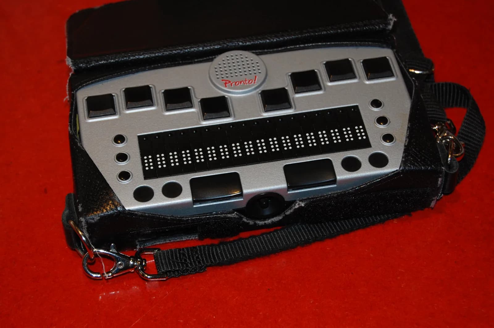 A portable Pronto braille notetaker with a row of 18 refreshable braille cells and eight input keys, in a black carrying case on a red surface