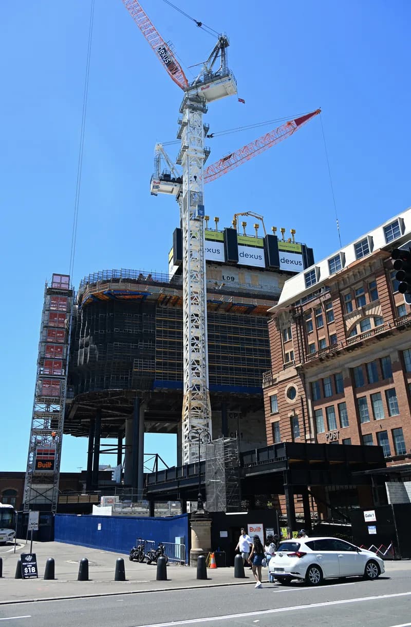 Tall building under construction with a tower crane beside a red-brick heritage building on a sunny Sydney street