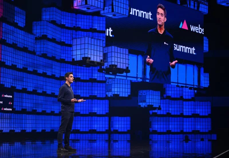 Feross Aboukhadijeh speaking on a stage lit in blue, with his image shown on a large screen behind him