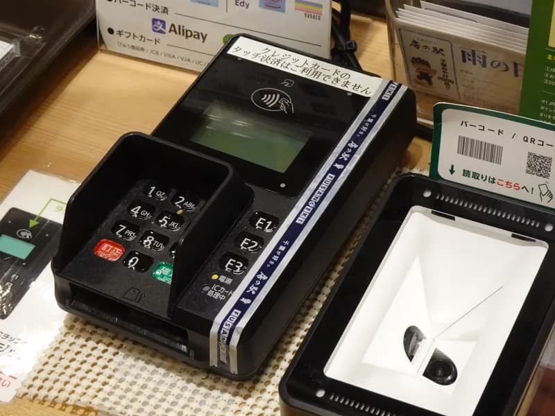Card payment terminal with keypad and contactless reader beside a QR code scanner on a shop counter in Japan