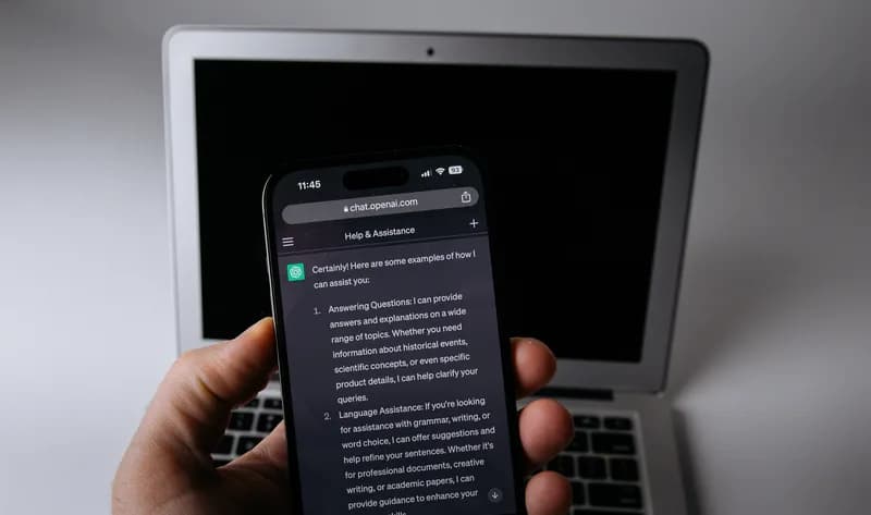 A hand holds a smartphone showing a ChatGPT conversation in front of an open laptop with a dark screen on a plain light background