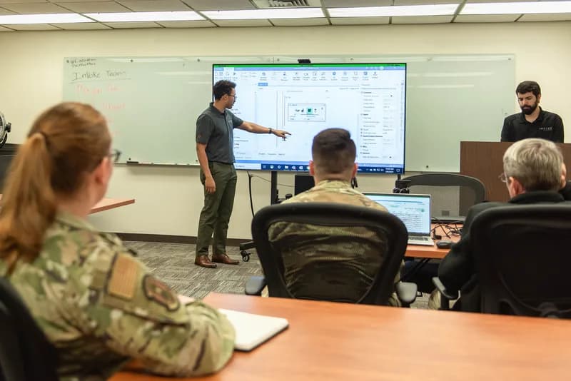 An instructor points at a large screen showing a UiPath automation workflow while service members and civilians follow along in a training room