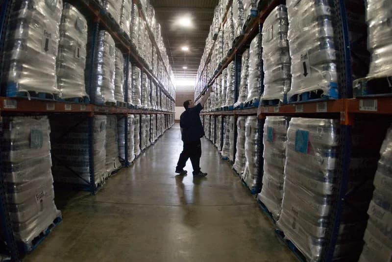 A worker in a long warehouse aisle reaches up to scan a barcode on shrink-wrapped pallets stacked on tall blue and orange racking