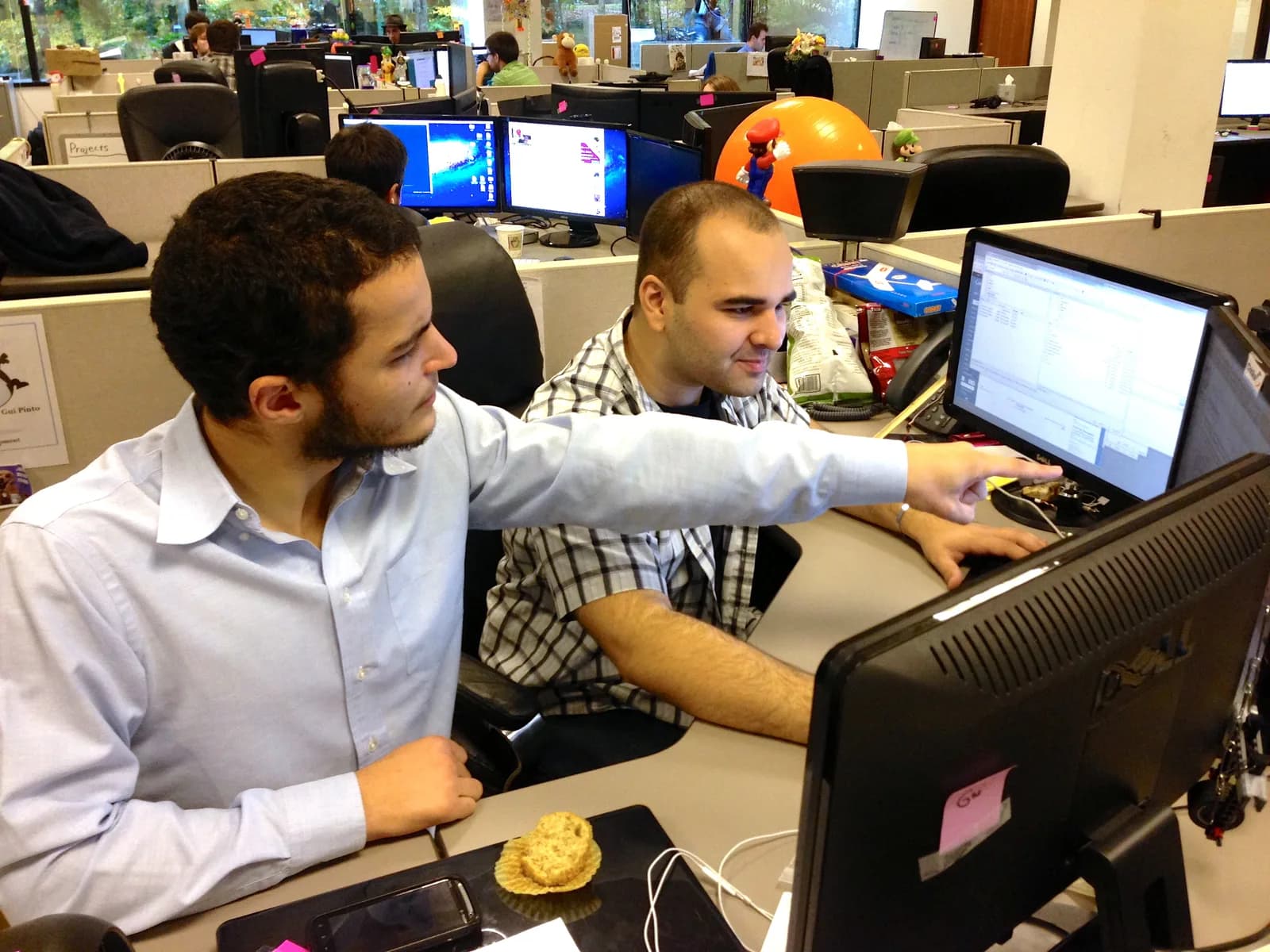 Two developers at a shared desk in an open-plan office, one pointing at code on a monitor while the other types, with more workstations behind them