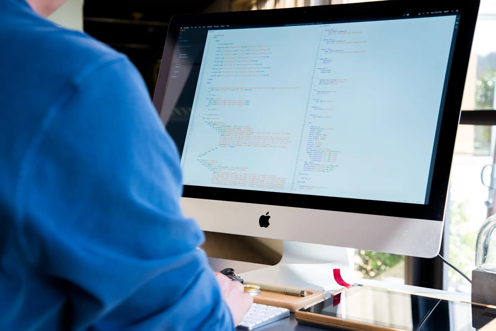 A developer in a blue shirt, seen from behind, writing code in a split-pane editor on an iMac at a bright desk