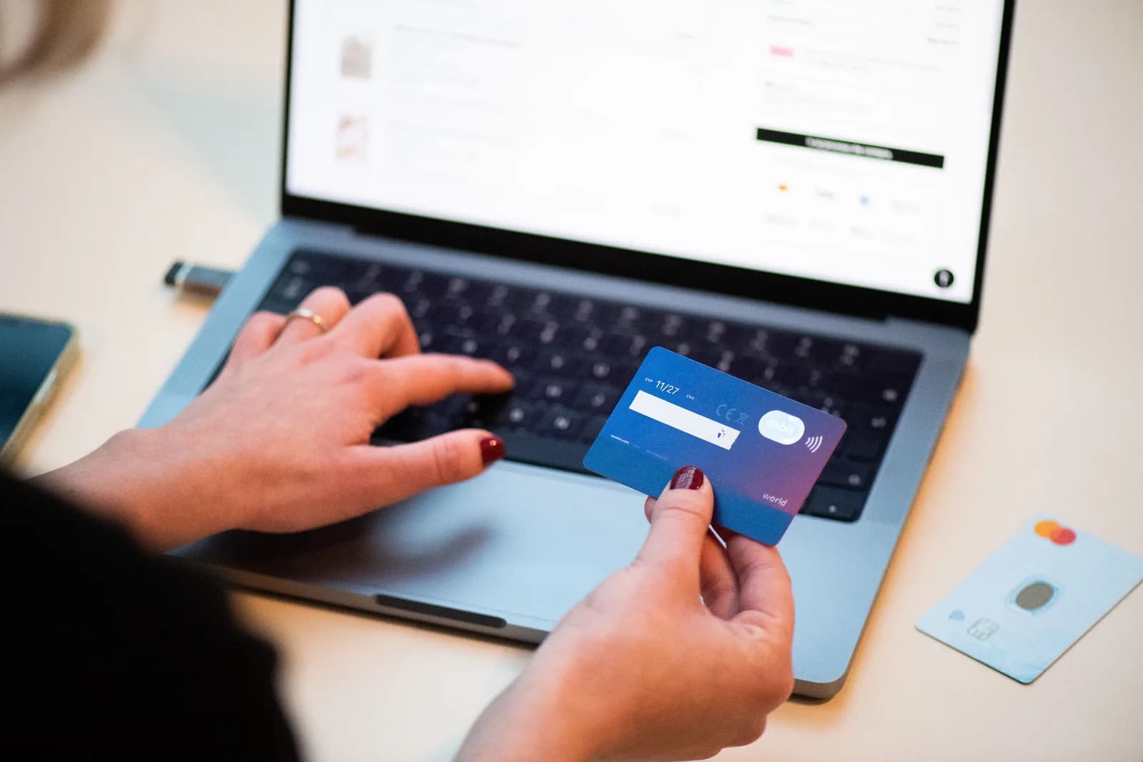 Close-up of a person holding a bank card while typing on a laptop that shows an online shop checkout page, with a second card on the desk