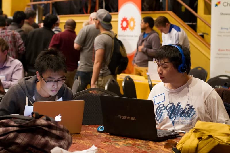 Two young developers working on laptops at a table during a TechCrunch hackathon in New York, with other attendees walking past behind them